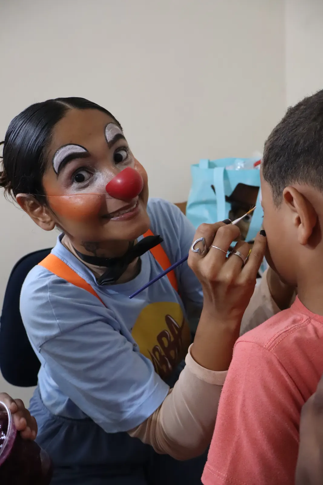 Menina com o rosto pintado de palhaço sendo retocada por uma voluntária